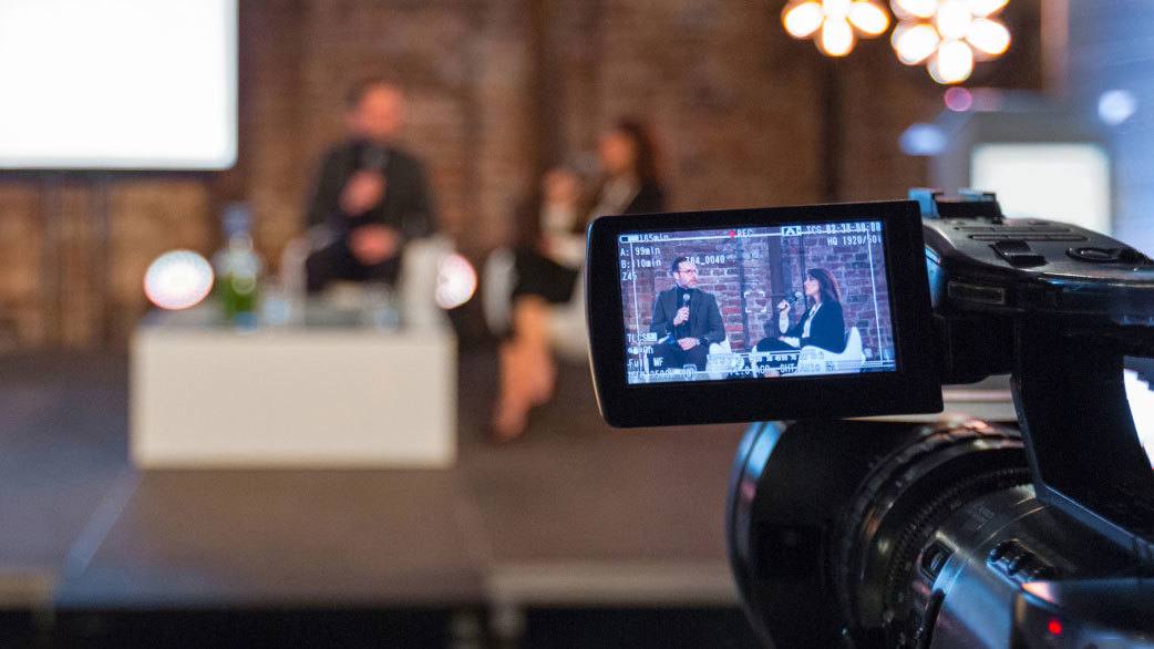 A blurry man and woman doing a tv interview in front of a brick wall with a video production camera in the foreground